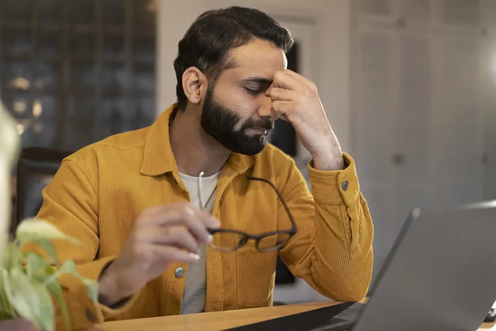 SEO techniques: A man in a mustard yellow corduroy shirt appears stressed as he sits at a desk, holding his glasses in one hand and massaging the bridge of his nose with the other, in front of a laptop, indicating a moment of frustration or headache while working.