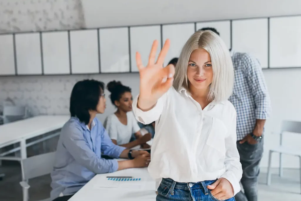 SEO techniques: A confident young woman with platinum blonde hair and wearing a white blouse and blue jeans extends her hand toward the camera in a high-five gesture after using white hat SEO techniques. In the softly lit background, three colleagues engage in discussion around a meeting table in a modern office setting.