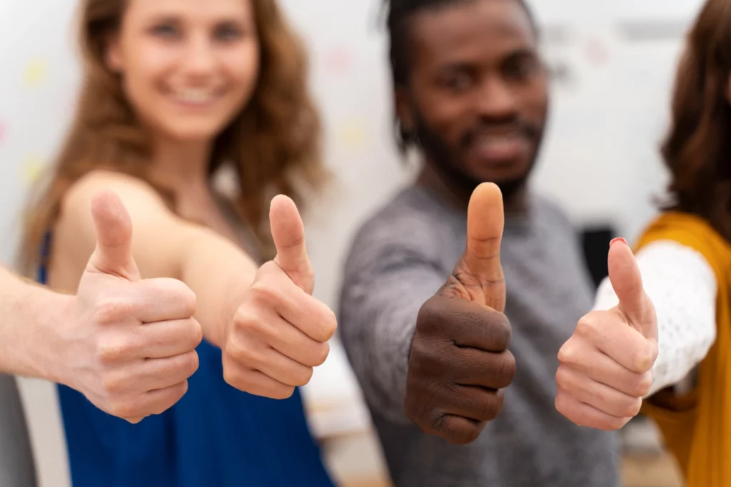 Group of diverse smiling people giving thumbs up in a bright collaborative workspace, indicating positive teamwork and success.