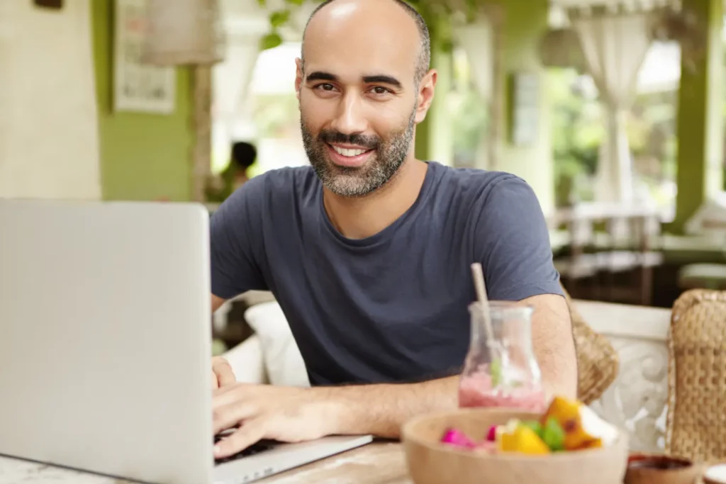 Smiling man using a laptop in a bright café with a refreshing drink and fruit bowl, illustrating the ease of browsing a website with optimized Core Web Vitals