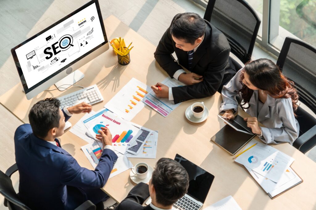 A diverse team of three business professionals collaboratively discussing over a laptop in an office setting, analyzing charts related to the pros and cons of SEO.