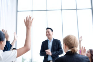 A presenter smiles in front of a group as audience members raise their hands, actively participating in the discussion, demonstrating strong audience engagement during the seminar.