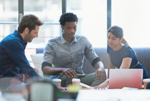 Three professionals sit together around a table, reviewing notes and a laptop, discussing strategies for their agency marketing services to enhance a client’s brand presence.