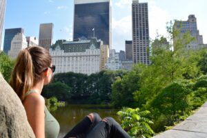 A woman sits on a rock, gazing at the skyline of a vibrant city, with tall buildings and lush greenery surrounding a calm pond. The serene scene reflects the blend of nature and urban life, a characteristic of cities like Vancouver, making it a prime location for a Google search company in Vancouver.