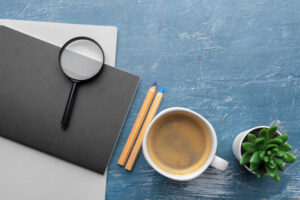 Top view of a modern workspace featuring a magnifying glass, two pencils, a coffee cup, and a small plant on a blue table. The setup symbolizes focus and precision, essential qualities in providing effective keyword research services.