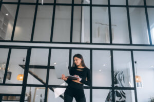 A young woman in a sports outfit standing confidently while holding a laptop in a modern, minimalistic space. The image represents the blend of fitness, lifestyle, and digital and social media marketing, showcasing how influencers and marketers use technology to engage their audiences online.