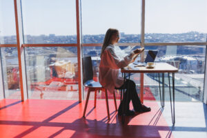 A professional woman wearing glasses, sitting in a brightly lit cafe with a panoramic view of Vancouver, working on a laptop and reviewing local search marketing materials while enjoying a coffee.