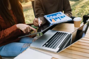 Two individuals working outdoors, discussing data from a tablet and a printed report with charts, alongside a laptop. This setting showcases teamwork and brainstorming digital marketing examples for business growth.
