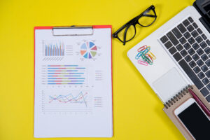 Top view of a workspace featuring a laptop, smartphone, notebook, and a clipboard displaying a colorful marketing report with charts and graphs, set against a bright yellow background.