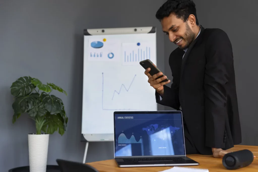 business-person-looking-finance-graphs Google ads ad scheduling benefits being assessed by a smiling business man holding a smart phone in an office with an open laptop and a display board depicting charts
